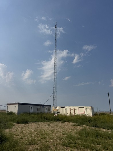 Lattice telecom tower rising above site containers and a field office building under an open Alberta prairie sky.