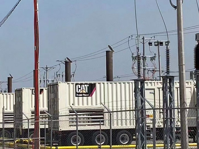 White containerized CAT Rental Power generator sets with exhaust stacks behind a fence, adjacent to overhead electrical distribution lines.